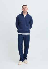 Felpa navy con zip e colletto alto, abbinata a pantaloni in denim blu e sneakers beige su uno sfondo neutro.