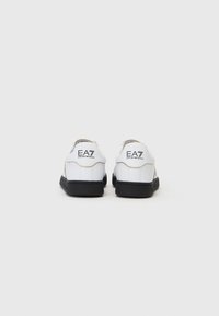 Sneakers bianchi con suole nere, caratterizzati da una texture in pelle liscia, dettagli dorati e il logo EA7 in nero sul tallone posteriore.