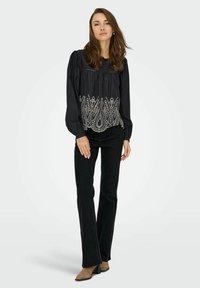 Blouse noire à manches longues avec épaules froncées et broderie blanche complexe sur l'ourlet, associée à un jean noir évasé et des bottines beige.