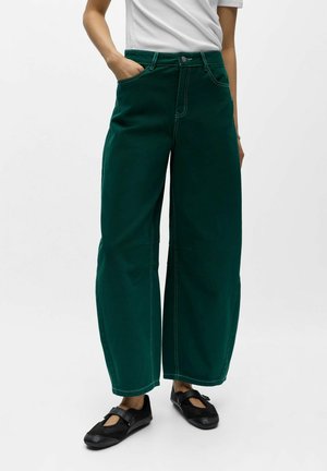 Personne portant un pantalon large vert foncé, un haut blanc et des chaussures Mary Jane noires, debout avec une main dans la poche.