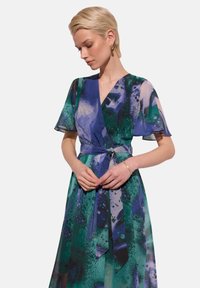 Sukienka w kolorze zielonym i niebieskim w abstrakcyjny wzór, z falbankowymi rękawami, paskiem w talii i przezroczystą teksturą.