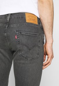 Jeans de mezclilla gris con un parche de cuero marrón en la cinturilla, que cuentan con un diseño distintivo en el bolsillo trasero y un detalle de etiqueta roja.