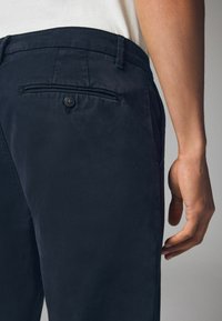 Pantaloni blu navy realizzati in un tessuto liscio, con una tasca posteriore con bottone e una vestibilità sartoriale, mostrati di lato con una mano nelle vicinanze.