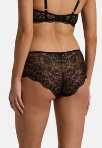 Ropa interior de encaje negro con patrones florales, bordes ondulados y una cinturilla suave. La vista trasera destaca los intrincados detalles de encaje.