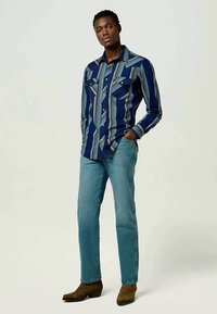 Camisa de mangas longas azul-marinho com riscas verticais coloridas, dois bolsos no peito e um colarinho pontiagudo, combinada com jeans azul claro e botas castanhas.