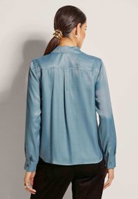 Lichtblauwe satijnen blouse, lange mouwen, rechte zoom, plooidetail achter, met knopen bij de manchetten, gladde textuur, gedragen met donkere broeken.