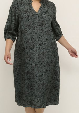 Woman wearing a loose, knee-length dark green dress with black abstract floral patterns and three-quarter puffed sleeves, standing with arms slightly extended.