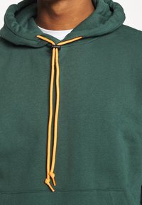 Felpa verde con cappuccio, caratterizzata da coulisse arancioni e un regolatore nero. Tessuto misto cotone, design casual.