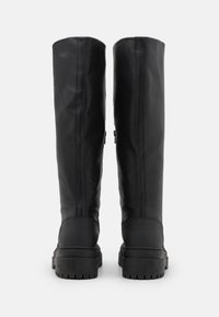 Korkeat mustat nahkasaappaat, joissa on paksut kumipohjat, näkymä takaa valkoista taustaa vasten.