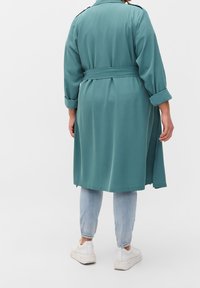 Persona che indossa un trench coat teal con cintura, jeans azzurri chiari con dettagli in strass sui polsini e sneakers bianche, mentre si trova contro uno sfondo neutro.