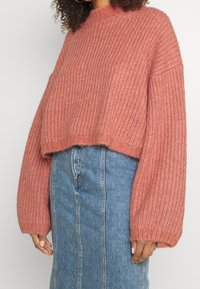 Kvinna i en lös rosa ribbad stickad tröja och en högmidjad blå denimkjol, med händerna avslappnat vid sidorna.