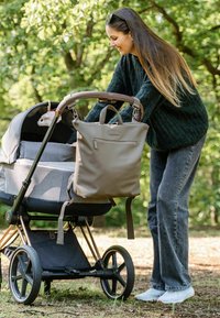 Harmaa lastenvaunu ruskealla kahvalla, jossa on kankaasta valmistettu katos ja taupe värinen säilytyslaukku, jossa on hihnat. Taustalla viherrystä.