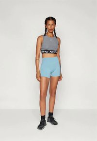 Top sport gri cu bandă "PRO NIKE", pantaloni scurți bleu deschis și adidași negri. Modelul stă în picioare cu brațele relaxate pe lângă corp.