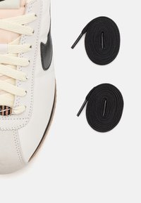 Vit sneaker med crèmefärgade snören och svart logotyp bredvid två rullade svarta skosnören på en vit bakgrund.