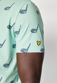 Mint green short-sleeve shirt with a golf club pattern in navy blue. Features a yellow embroidered logo on the sleeve. Smooth texture.