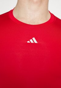 Red athletic shirt made of smooth fabric with a crew neckline and white adidas logo on the chest. Fitted shape, minimalistic design.