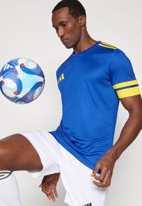 Maglia da atletica blu con strisce gialle sulle spalle, abbinata a pantaloncini bianchi. Il giocatore tiene in equilibrio una palla da calcio multicolore vicino al ginocchio.