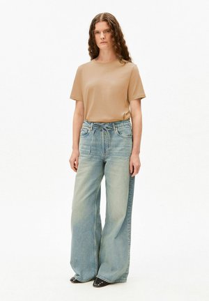 Jong persoon met krullend bruin haar die een beige shirt met korte mouwen draagt en losse lichtblauwe high-waisted wijde jeans met zwarte schoenen met studs.
