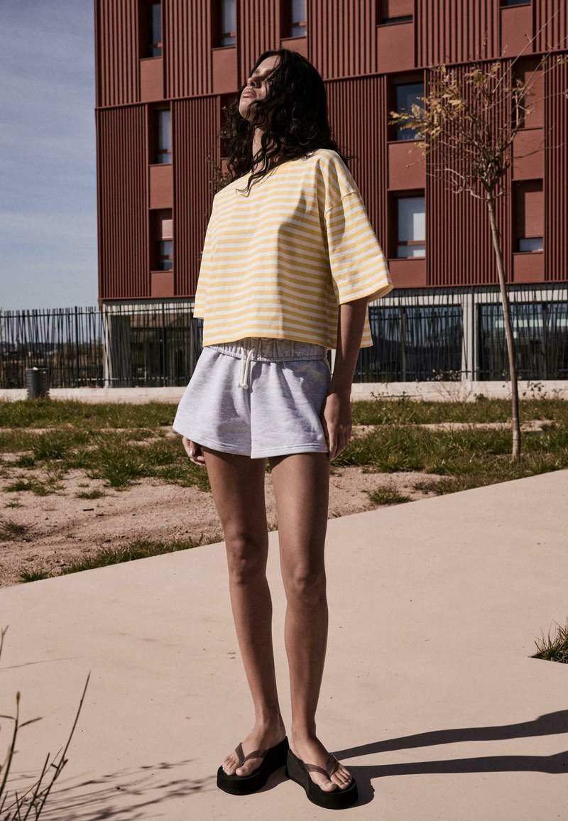 Jeune personne aux cheveux bouclés porte un t-shirt à rayures jaunes, un short gris et des tongs noires à plateforme, debout à l'extérieur près d'un bâtiment rouge.
