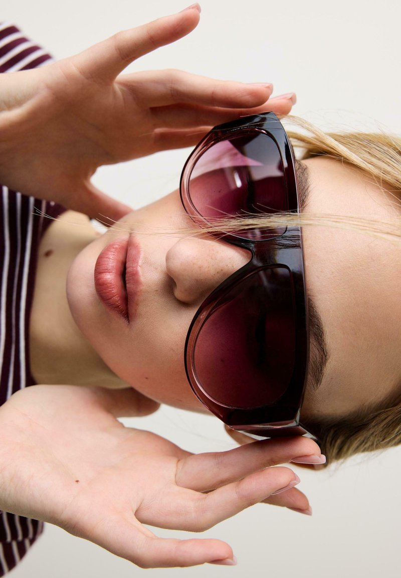 Personne blonde ajustant de grandes lunettes de soleil rouge foncé, les lèvres légèrement entrouvertes et portant un haut rayé sur un fond uni.
