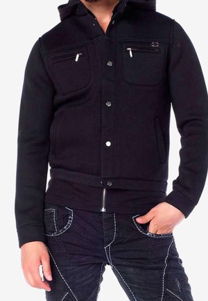 Mann trägt einen schwarzen Hoodie mit Reißverschluss, der mit Knöpfen versehene Taschen und dunkelblaue Jeans mit kontrastierenden weißen Nähten hat, eine Hand in der Tasche.