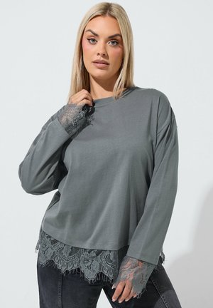 Blondynka ubrana w luźny szary top z długim rękawem ozdobiony koronką na rękawach i dole, w połączeniu z ciemnymi spodniami, na jednolitym tle.