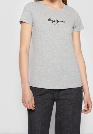 Kvinde iført en lysegrå Pepe Jeans London t-shirt og mørkegrå jeans står mod en ensfarvet lys baggrund.