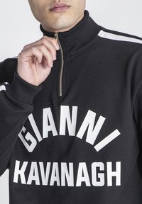 Čierna mikina na zips s vysokým golierom, s odvážnym bielym textom "GIANNI KAVANAGH." Textília vyzerá hladko s rebrovanými manžetami.