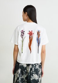 Hvit bomull t-skjorte med akvarell blomsterdesign i lilla, rød og blå på baksiden. Korte ermer og løs passform.