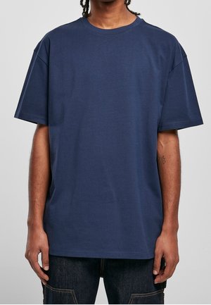 Mann trägt ein schlichtes navyblaues, übergroßes T-Shirt mit kurzen Ärmeln und dunkle Jeans, steht vor einem schlichten hellgrauen Hintergrund.