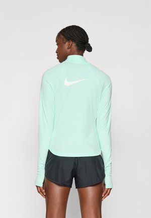 Mintgrønn langermet idrettstopp med høy krage og hvit Nike-logo på øvre rygg, sammen med svarte shorts. Jevn tekstur.