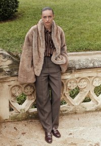 Žena stoji na otvorenom pored kamenog rukohvata, nosi bež kaput od krzna, smeđu prugastu košulju, široke smeđe hlače i smeđe mokasine.