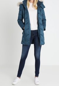 Tyrkis pufferjakke med pelsforet hætte, quiltet design, to frontlommer, sammen med mørke jeans og hvide sneakers.