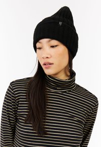 Bonnet tricoté noir avec un sommet pointu, présentant une texture côtelée et une petite étiquette. Le modèle porte un col roulé rayé noir et doré.