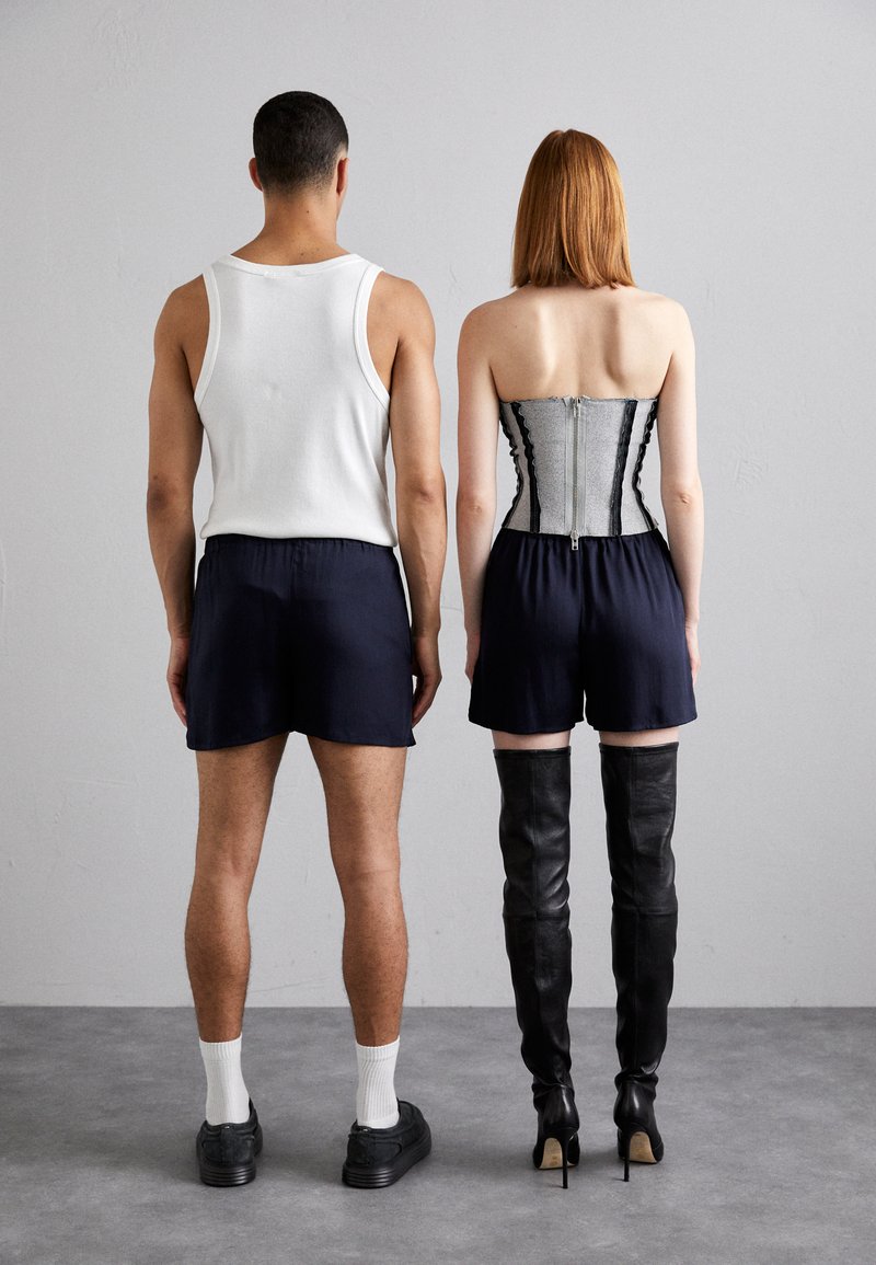 Ein Paar von Personen, die sich abgewandt haben. Eine trägt ein weißes Tanktop und eine marineblaue Shorts, die andere trägt ein silbernes Korsett, marineblaue Shorts und kniehohe schwarze Stiefel.