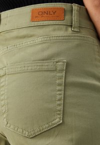 Olivgröna byxor med en texturerad yta, försedd med ett brunt läderlogotypmärke, två bakfickor och karakteristiska sömdetaljer.