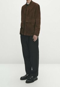 Bruine suÈde jas met ritssluiting en twee voorzakken, gecombineerd met losse zwarte broek en zwarte veterschoenen.