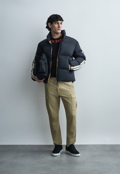 Ung mand iført sort dunjakke med hvide striber, beige cargobukser, sorte sneakers, holder en sort skuldertaske, stående mod en ensfarvet væg.