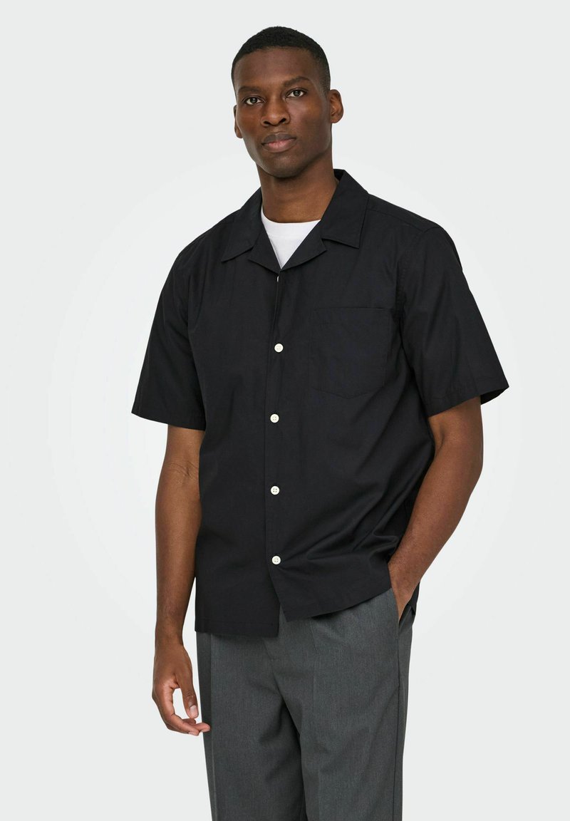 Homme portant une chemise noire à manches courtes boutonnée par-dessus un t-shirt blanc et un pantalon gris, debout avec une main dans la poche devant un fond uni.