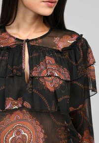 Blouse noire transparente avec un col froncé, des motifs floraux complexes en orange et rose, et une ouverture en forme de cœur fermée par un bouton.