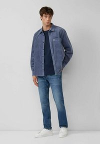 Junger Mann steht gegen eine Wand und trägt ein blaues Cordhemd, ein marineblaues T-Shirt, blaue Jeans und weiße Sneakers.