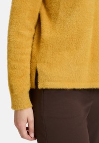 Żółty, puszysty sweter o teksturowanym wykończeniu, z bocznymi rozcięciami, noszony z dopasowanymi ciemnobrązowymi spodniami. Zbliżenie na detale rękawa i krawędzi.