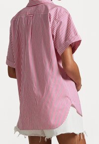 Camicia a maniche corte a righe rosa e bianche con spacco posteriore, orlo curvo e vestibilità rilassata. Il materiale sembra leggero; la texture è liscia.