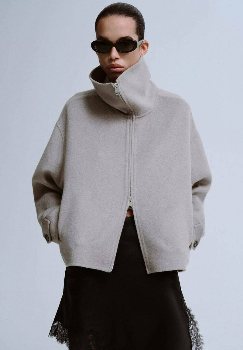 Lichtgrijze oversized jas met hoge kraag, ritssluiting aan de voorkant en een losse pasvorm. Gedragen over een zwarte satijnen rok met kanten rand en donkere zonnebril.