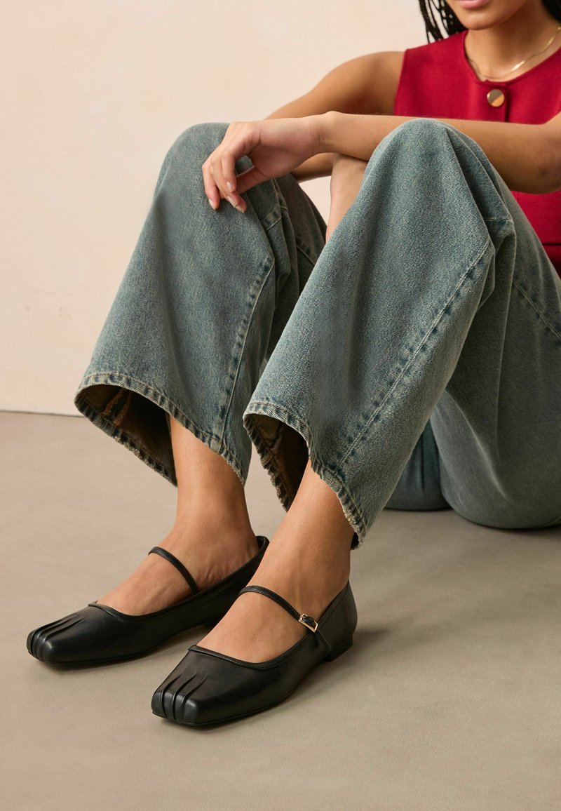 Black leather flats with a unique toe design, adjustable ankle strap, and a low heel. Worn with wide-leg denim jeans.