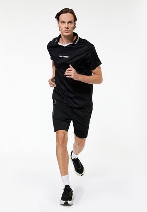 Hombre corriendo hacia adelante con pantalones cortos deportivos negros, camiseta negra con texto "TRUST YOURSELF", calcetines blancos y zapatillas negras sobre fondo blanco.