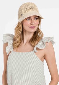 Sombrero beige similar a la paja, con una parte superior redondeada y un ala ancha y curva. Presenta un pequeño acento plateado y un diseño de tejido abierto texturizado.