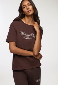 Brązowa bawełniana koszulka z krótkim rękawem, z białym napisem "Athleisure Club" oraz "Relaks i Wellness". Model pozuje z rękami skrzyżowanymi.