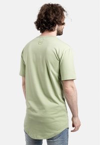 Camiseta de algodón verde claro con mangas cortas, dobladillo curvado y un pequeño logo bordado en la parte posterior cerca del cuello. Llevada con jeans azules.