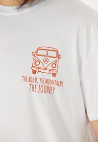 Vit t-shirt med en orange grafik av en vintage furgon och texten "VÄGEN, BERGEN, RESAN." Mjuk tyg.